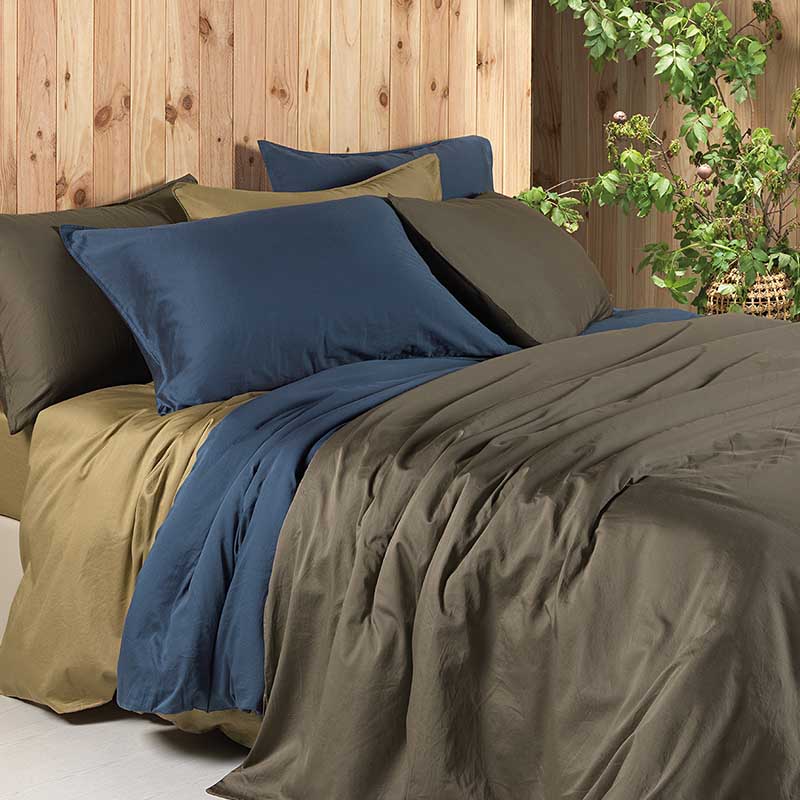 planken muur, plant en bed met drie dekbedden en kussens in blauw, leem en brons